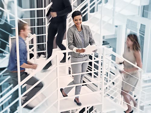 Young professional woman standing on in the middle of a stairway with colleagues ascending and descending around her.