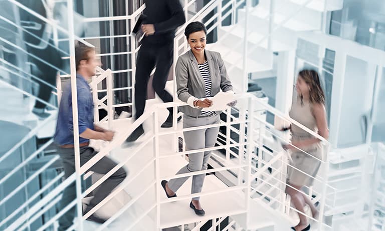 Young professional woman standing on in the middle of a stairway with colleagues ascending and descending around her.